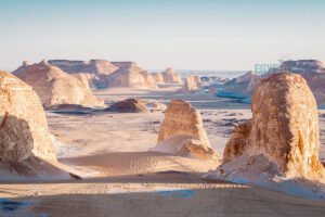 A wide-angle landscape photograph capturing massive, unique wind-eroded rock formations and mesas rising from the desert floor under a clear sky in the Egyptian Western Desert.
