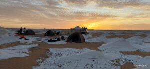 A wide-angle landscape photograph of a desert base camp at dawn, with four dark expedition tents on a mixed surface of sand and pale, salt-crusted earth. Several people gather around a cooking fire, and a conical peak is visible on the distant horizon.