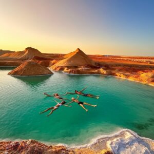 Tourists floating effortlessly in a vibrant turquoise salt lake surrounded by golden sand dunes during Private desert tours Egypt.