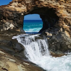 A natural rock arch framing a view of the bright blue ocean, with a foamy waterfall cascading over rugged brown rocks in the foreground.